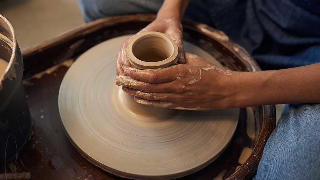A student working clay into a hollow cylinder on a spinning wheel