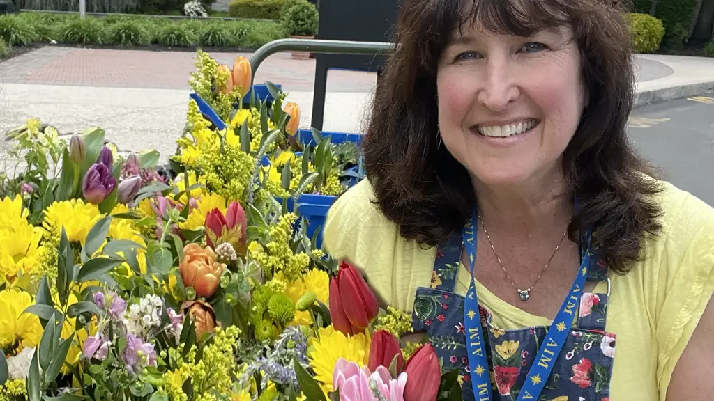 Sharon Freed Mosenkis, workshop director for the art center's Mother's Day floral arrangement workshop, smiles behind a cart overflowing with an abundant display of fresh-cut flowers and foliage. She is surrounded by colorful tulips, yellow solidago, sweet peas, and assorted leaves stacked in blue crates. Sharon wears a yellow top and an apron with a blue 'AIM' lanyard. Background: Paved exterior of an art center.
